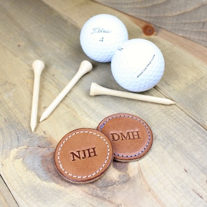 Puede incluir: Dos marcadores de bola de golf de cuero marrón con las iniciales "DMH" y "NJH" grabadas en ellos. Los marcadores están sobre una superficie de madera con dos bolas de golf y tres tees.