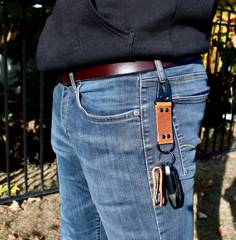 May include: A brown leather key chain with a black metal clip and two keys attached. The key chain is attached to a belt loop on a pair of blue jeans.