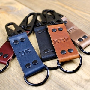 May include: Five leather keychains with black metal rings and clasps. Each keychain has a different color and is personalized with initials. The initials are "DK", "KRF", and "NIL".