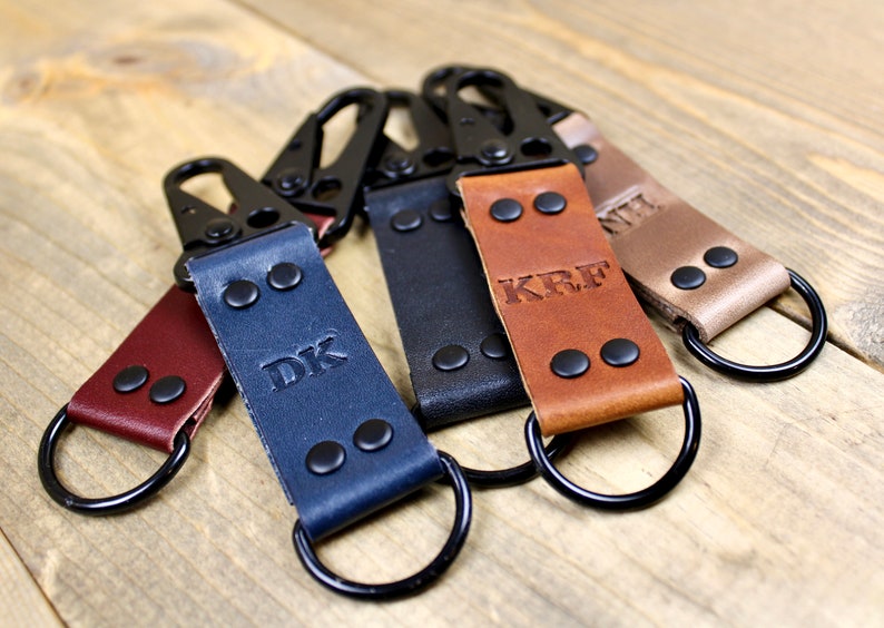 May include: Five leather keychains with black metal rings and clasps. Each keychain has a different color and is personalized with initials. The initials are "DK", "KRF", and "NIL".
