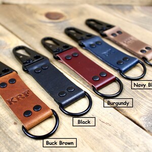 May include: Five leather keychains with black metal clasps and rings. The keychains are in the colors buck brown, black, burgundy, navy blue, and brown.