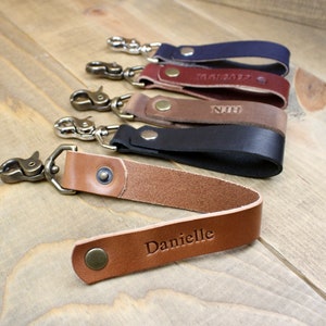 May include: Five leather keychains with metal clasps and personalised engravings. The keychains are brown, black, and navy blue. The engravings include "Danielle", "NH", "01/01/2022", and "MAI 2022".