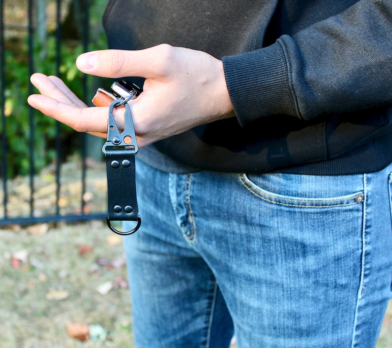 May include: A black leather key chain with a metal clip and a ring. The key chain is attached to a pair of keys.
