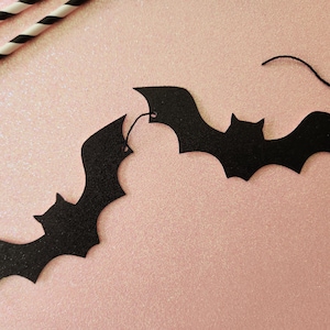 May include: Black bat-shaped garland with a string for hanging, perfect for Halloween decorations. The bats are made of a textured material and are set against a pink background. Two black and white striped paper straws are in the upper left corner.