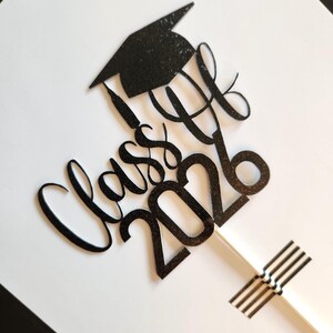 May include: A black glittery cake topper featuring a graduation cap icon above the cursive text "Class of 2026". The design is attached to a thin white stick, which is secured at the bottom with a small white rectangular piece for stability.