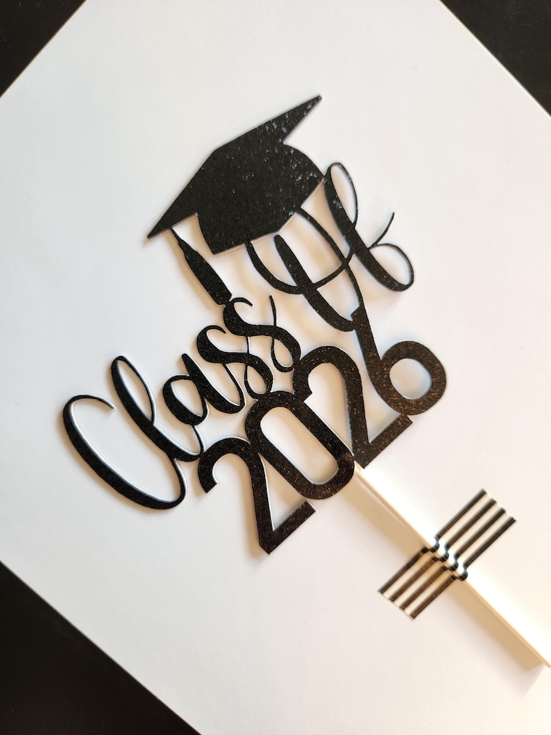 May include: A black glittery cake topper featuring a graduation cap icon above the cursive text "Class of 2026". The design is attached to a thin white stick, which is secured at the bottom with a small white rectangular piece for stability.