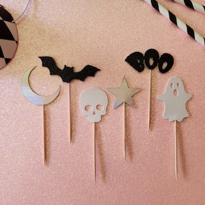 May include: Halloween-themed cupcake toppers in various shapes, including a black bat, a silver crescent moon, a skull, a silver star, and a white ghost. The word "BOO" is spelled out in black felt. The toppers are on wooden sticks, set against a pink glitter background.