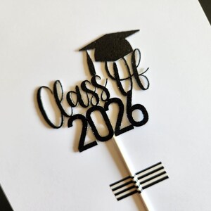 May include: A black glittery cake topper featuring the text Class of 2026 in a cursive script font with a graduation cap icon positioned above the text. The topper is attached to a thin white stick, which is secured with black and white striped tape.