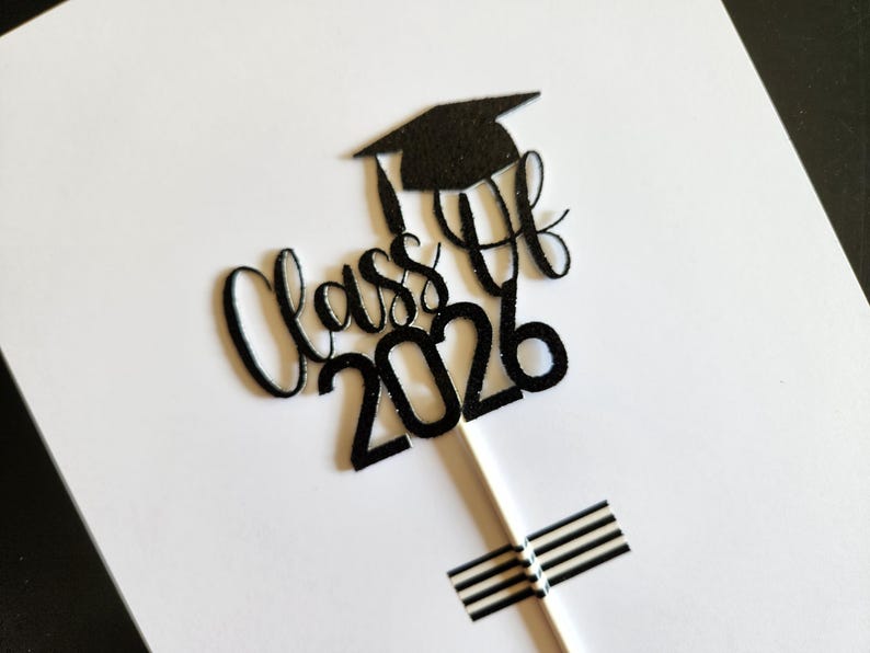 May include: A black glittery cake topper featuring the text Class of 2026 in a cursive script font with a graduation cap icon positioned above the text. The topper is attached to a thin white stick, which is secured with black and white striped tape.
