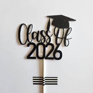 May include: A black glittery cake topper featuring the text Class of 2026 in a cursive and block font combination. A graduation cap icon sits above the text. The topper is attached to a thin white stick, which is secured by a piece of black and white striped tape.