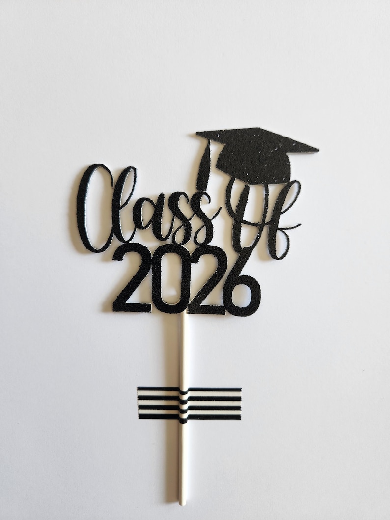 May include: A black glittery cake topper featuring the text Class of 2026 in a cursive and block font combination. A graduation cap icon sits above the text. The topper is attached to a thin white stick, which is secured by a piece of black and white striped tape.