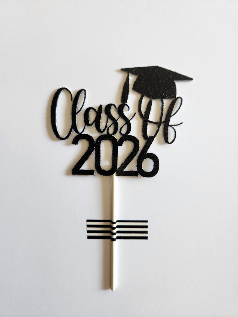 May include: A black glittery cake topper featuring the text "Class Of 2026" in a cursive script font, topped with a graduation cap icon. The topper is attached to a thin white stick, which is secured by a small piece of black and white striped tape.
