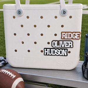 May include: A white Bogg tote bag with a perforated body and two handles. The bag features three rectangular name tags attached to the front, displaying the names RIDGE, OLIVER, and HUDSON in white block lettering on brown, grey, and black backgrounds.