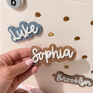 Puede incluir: Un bolso de color crema con superficie perforada presenta inserciones de nombres personalizables. El bolso muestra etiquetas de nombres tridimensionales que dicen Luke, Sophia y Brooklyn en varios colores y texturas, incluyendo brillo.