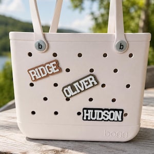 May include: A light pink tote bag with two handles and a perforated body featuring the embossed text bogg at the bottom. Three rectangular name charms are attached to the holes, displaying the names RIDGE in brown, OLIVER in olive, and HUDSON in black.