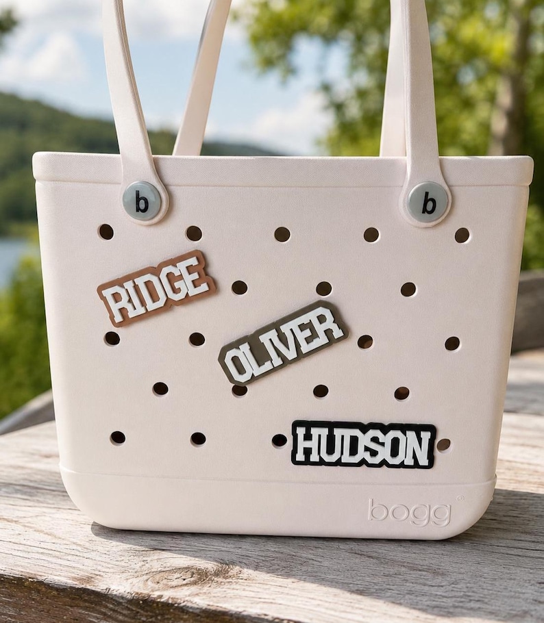 May include: A light pink tote bag with two handles and a perforated body featuring the embossed text bogg at the bottom. Three rectangular name charms are attached to the holes, displaying the names RIDGE in brown, OLIVER in olive, and HUDSON in black.