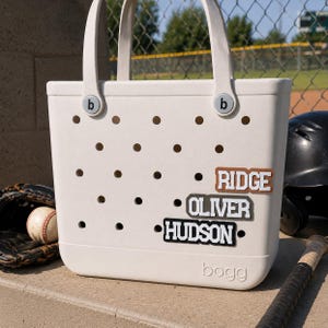 May include: A cream-colored Bogg tote bag with a perforated body and two handles. The bag features three rectangular name tags attached to the front, displaying the names RIDGE in brown, OLIVER in gray, and HUDSON in black with white lettering.
