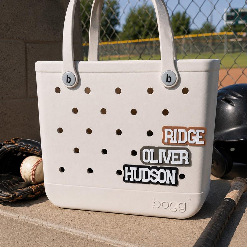 May include: A cream-colored Bogg tote bag with a perforated body and two handles. The bag features three rectangular name tags attached to the front, displaying the names RIDGE in brown, OLIVER in gray, and HUDSON in black with white lettering.