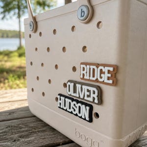 May include: A beige tote bag with a perforated surface and two handles. The bag features three rectangular name tags attached to the front, displaying the names RIDGE, OLIVER, and HUDSON in white lettering. The word bogg is embossed at the bottom.