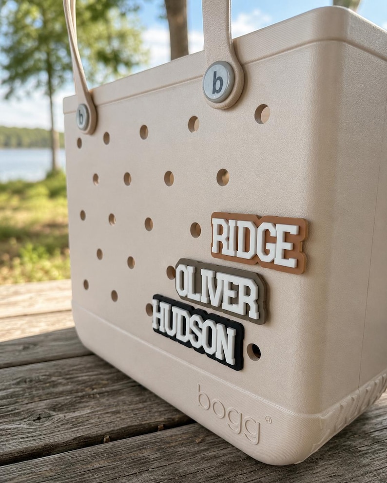 May include: A beige tote bag with a perforated surface and two handles. The bag features three rectangular name tags attached to the front, displaying the names RIDGE, OLIVER, and HUDSON in white lettering. The word bogg is embossed at the bottom.