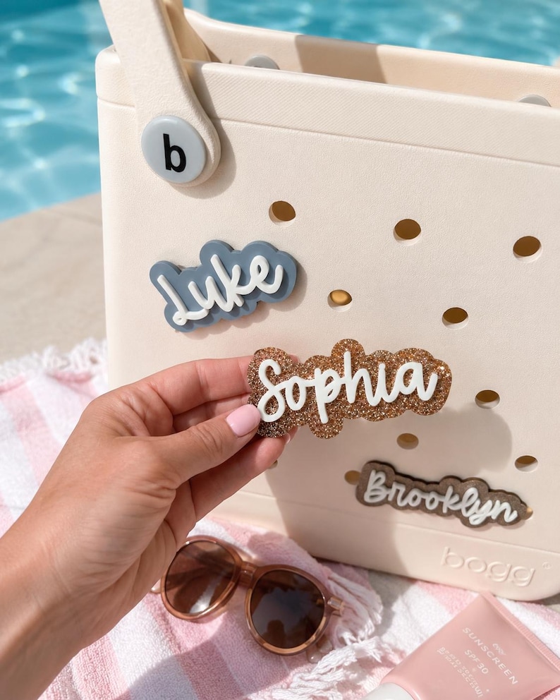 Puede incluir: Un bolso de color crema con superficie perforada presenta inserciones de nombres personalizables. El bolso muestra etiquetas de nombres tridimensionales que dicen Luke, Sophia y Brooklyn en varios colores y texturas, incluyendo brillo.