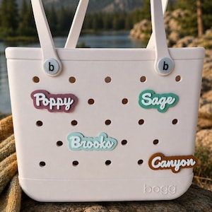 Puede incluir: Un bolso tote perforado de color rosa claro con dos asas y la palabra bogg grabada en la parte inferior. Cuatro inserciones de nombres personalizadas est&aacute;n fijadas en los agujeros, con los nombres Poppy en granate, Sage en verde azulado, Brooks en azul claro y Canyon en marr&oacute;n.