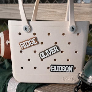 May include: A cream-colored tote bag with a perforated surface and two handles. The bag features the brand name Bogg embossed at the bottom and two circular b-logo buttons. Three rectangular name charms are attached, reading RIDGE, OLIVER, and HUDSON.