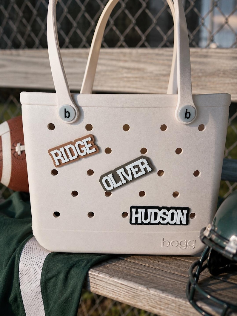 May include: A cream-colored tote bag with a perforated surface and two handles. The bag features the brand name Bogg embossed at the bottom and two circular b-logo buttons. Three rectangular name charms are attached, reading RIDGE, OLIVER, and HUDSON.