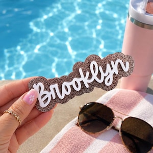 Puede incluir: Un letrero decorativo con la palabra Brooklyn en letras cursivas blancas sobre un fondo marr&oacute;n brillante con un borde festoneado. El letrero se sostiene frente a una piscina, un vaso rosa y unas gafas de sol.