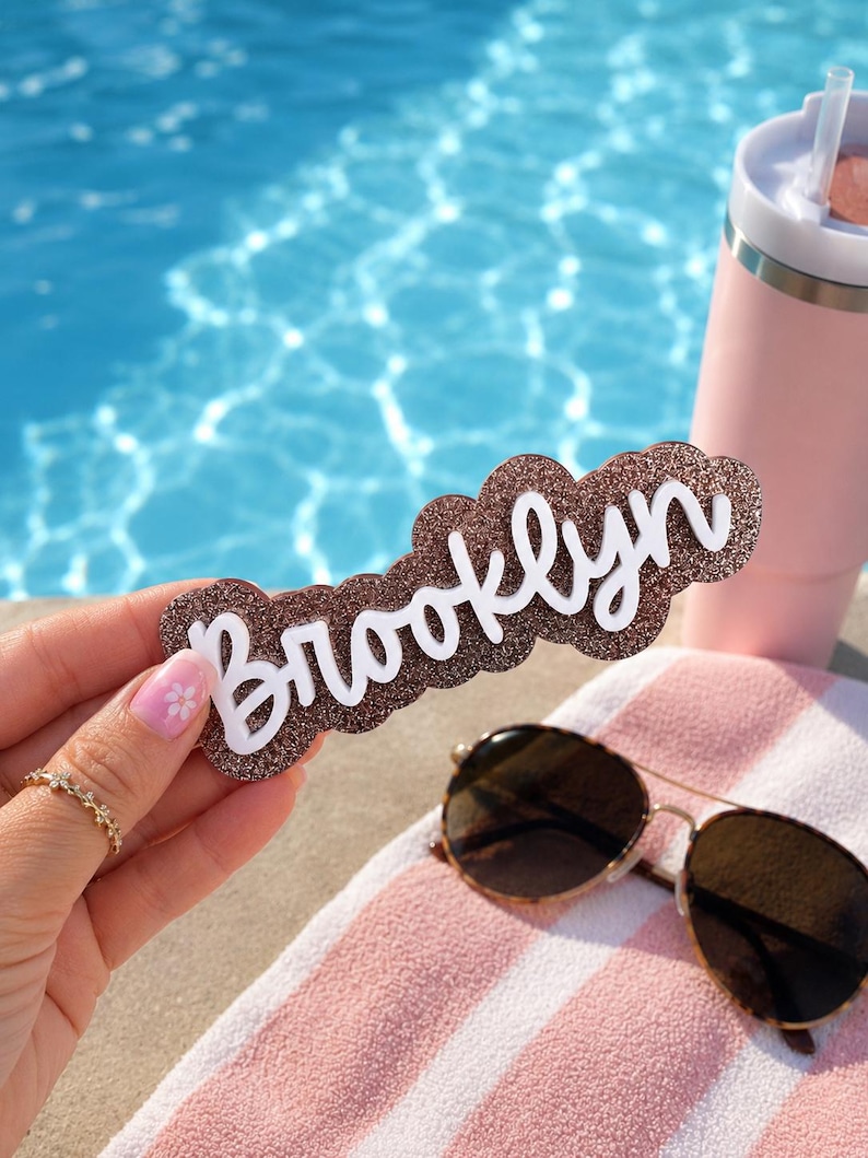 Puede incluir: Un letrero decorativo con la palabra Brooklyn en letras cursivas blancas sobre un fondo marr&oacute;n brillante con un borde festoneado. El letrero se sostiene frente a una piscina, un vaso rosa y unas gafas de sol.