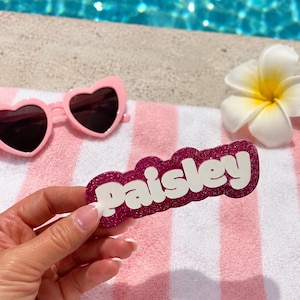 Puede incluir: Una etiqueta personalizada con el nombre Paisley en letras 3D blancas sobre un fondo festoneado de color rosa oscuro con purpurina. El art&iacute;culo se muestra junto a unas gafas de sol rosas en forma de coraz&oacute;n y una flor blanca sobre una toalla a rayas.