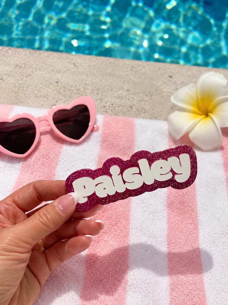 Puede incluir: Una etiqueta personalizada con el nombre Paisley en letras 3D blancas sobre un fondo festoneado de color rosa oscuro con purpurina. El art&iacute;culo se muestra junto a unas gafas de sol rosas en forma de coraz&oacute;n y una flor blanca sobre una toalla a rayas.