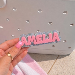 Puede incluir: Una etiqueta personalizada con el nombre AMELIA en letras may&uacute;sculas de color rosa mate en relieve sobre un fondo festoneado de purpurina plateada. La etiqueta est&aacute; dise&ntilde;ada para fijarse en los agujeros de un bolso blanco perforado.