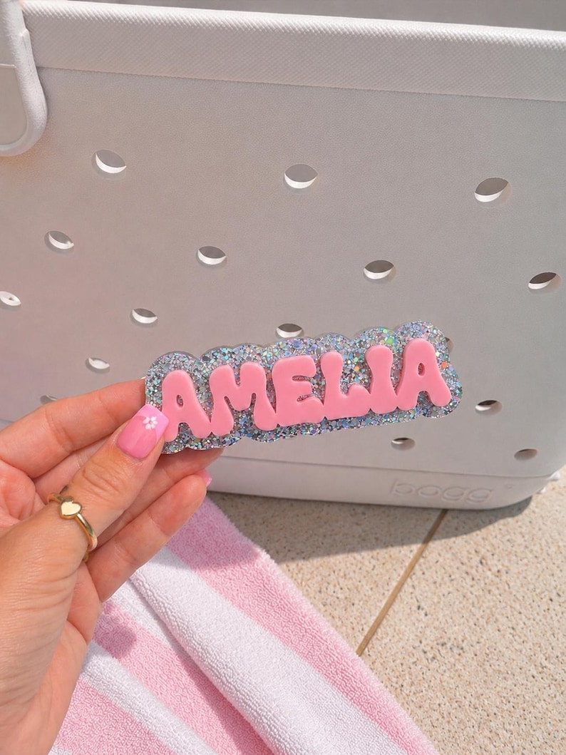 Puede incluir: Una etiqueta personalizada con el nombre AMELIA en letras may&uacute;sculas de color rosa mate en relieve sobre un fondo festoneado de purpurina plateada. La etiqueta est&aacute; dise&ntilde;ada para fijarse en los agujeros de un bolso blanco perforado.