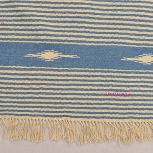 May include: A blue and cream-colored striped rug with a fringed edge. The rug features a central blue band with two cream-colored, stylized sunburst designs. The rug's design is composed of horizontal stripes.