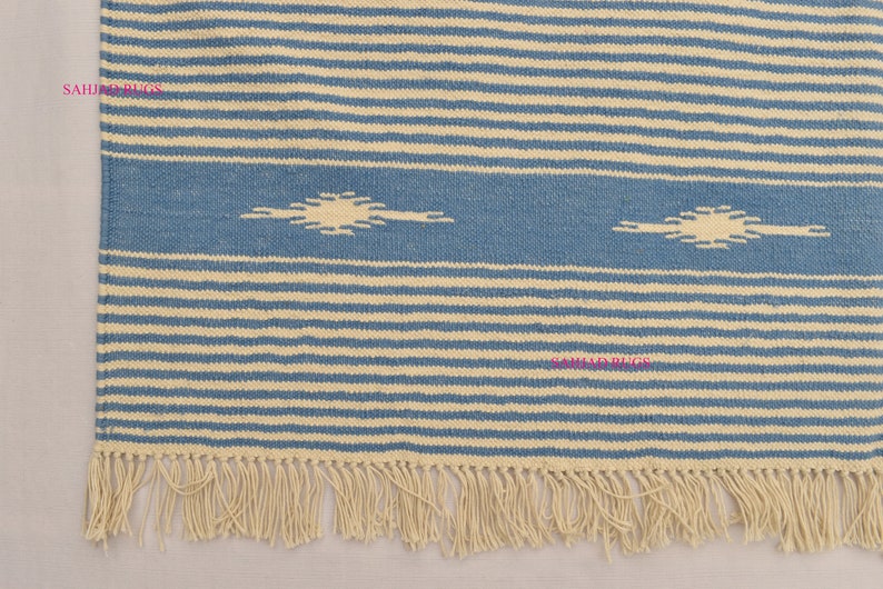 May include: A blue and cream-colored striped rug with a fringed edge. The rug features a central blue band with two cream-colored, stylized sunburst designs. The rug's design is composed of horizontal stripes.