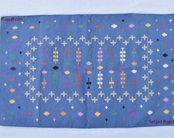 Könnte beinhalten: Ein blauer Teppich mit einem geometrischen Muster in Weiß, Schwarz und verschiedenen Farben. Der Teppich hat einen Fransenrand und der Text "Sahjad Rugs@com" ist in der oberen linken und unteren rechten Ecke sichtbar.