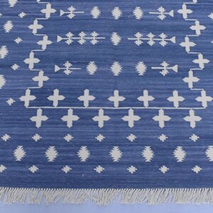 Puede incluir: Una alfombra azul y blanca con un borde de flecos. La alfombra tiene un dise&ntilde;o geom&eacute;trico con cruces y diamantes blancos sobre un fondo azul.