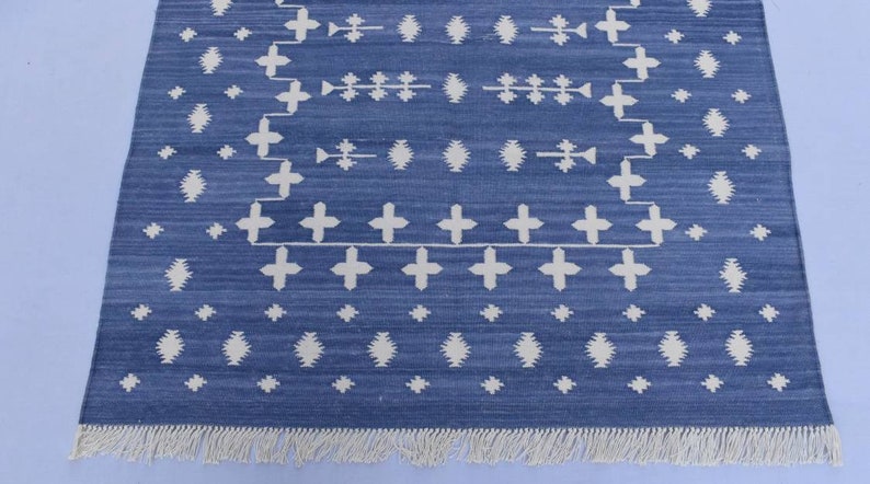 Puede incluir: Una alfombra azul y blanca con un borde de flecos. La alfombra tiene un dise&ntilde;o geom&eacute;trico con cruces y diamantes blancos sobre un fondo azul.