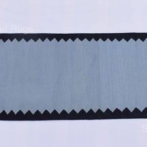 May include: A rectangular rug with a light blue center and a black zigzag border. The rug has a fringe on both ends. The rug is a decorative item for a home.