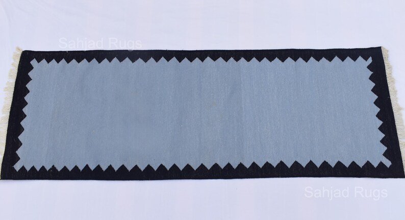 May include: A rectangular rug with a light blue center and a black zigzag border. The rug has a fringe on both ends. The rug is a decorative item for a home.