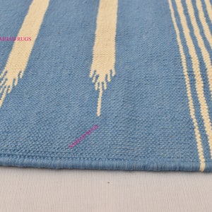 May include: A blue and white striped rug with a textured weave. The rug features wide, light blue stripes and off-white vertical stripes with a stylized design. The rug's edge is neatly finished with a blue border. The text "SAHJAD RUGS" is visible.
