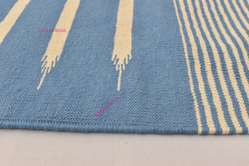 May include: A blue and white striped rug with a textured weave. The rug features wide, light blue stripes and off-white vertical stripes with a stylized design. The rug's edge is neatly finished with a blue border. The text "SAHJAD RUGS" is visible.