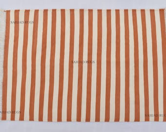 Könnte beinhalten: Ein gestreifter Teppich mit orangefarbenen und weißen Streifen. Der Teppich hat einen Fransenrand und der Text "SAHJAD RUGS" ist auf den Teppich gedruckt.