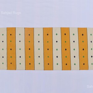 Puede incluir: Una alfombra rectangular con rayas verticales amarillas y blanquecinas. Peque&ntilde;os rombos verdes y azul oscuro est&aacute;n espaciados uniformemente a lo largo de las rayas. La alfombra tiene bordes con flecos y el texto "Sahjad Rugs" es visible.