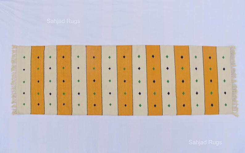 Puede incluir: Una alfombra rectangular con rayas verticales amarillas y blanquecinas. Peque&ntilde;os rombos verdes y azul oscuro est&aacute;n espaciados uniformemente a lo largo de las rayas. La alfombra tiene bordes con flecos y el texto "Sahjad Rugs" es visible.