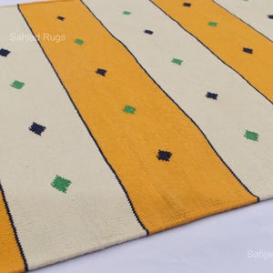 Puede incluir: Primer plano de una alfombra con rayas amarillas y blanquecinas alternas. Peque&ntilde;os acentos en forma de diamante azul oscuro y verde est&aacute;n dispersos por la alfombra. La alfombra tiene un aspecto texturizado y probablemente est&aacute; hecha de material tejido.
