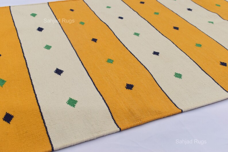Puede incluir: Primer plano de una alfombra con rayas amarillas y blanquecinas alternas. Peque&ntilde;os acentos en forma de diamante azul oscuro y verde est&aacute;n dispersos por la alfombra. La alfombra tiene un aspecto texturizado y probablemente est&aacute; hecha de material tejido.