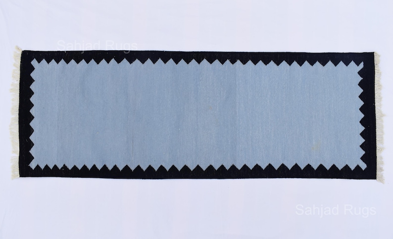 May include: Rectangular rug with a light blue center and a black zigzag border. The rug has a cream-colored fringe on the short ends. The rug is displayed on a white background. The rug is a home decor item.
