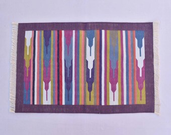 Puede incluir: Una alfombra rayada colorida con un patrón geométrico en tonos de morado, rosa, azul, amarillo y blanco. La alfombra tiene un borde morado y flecos blancos.